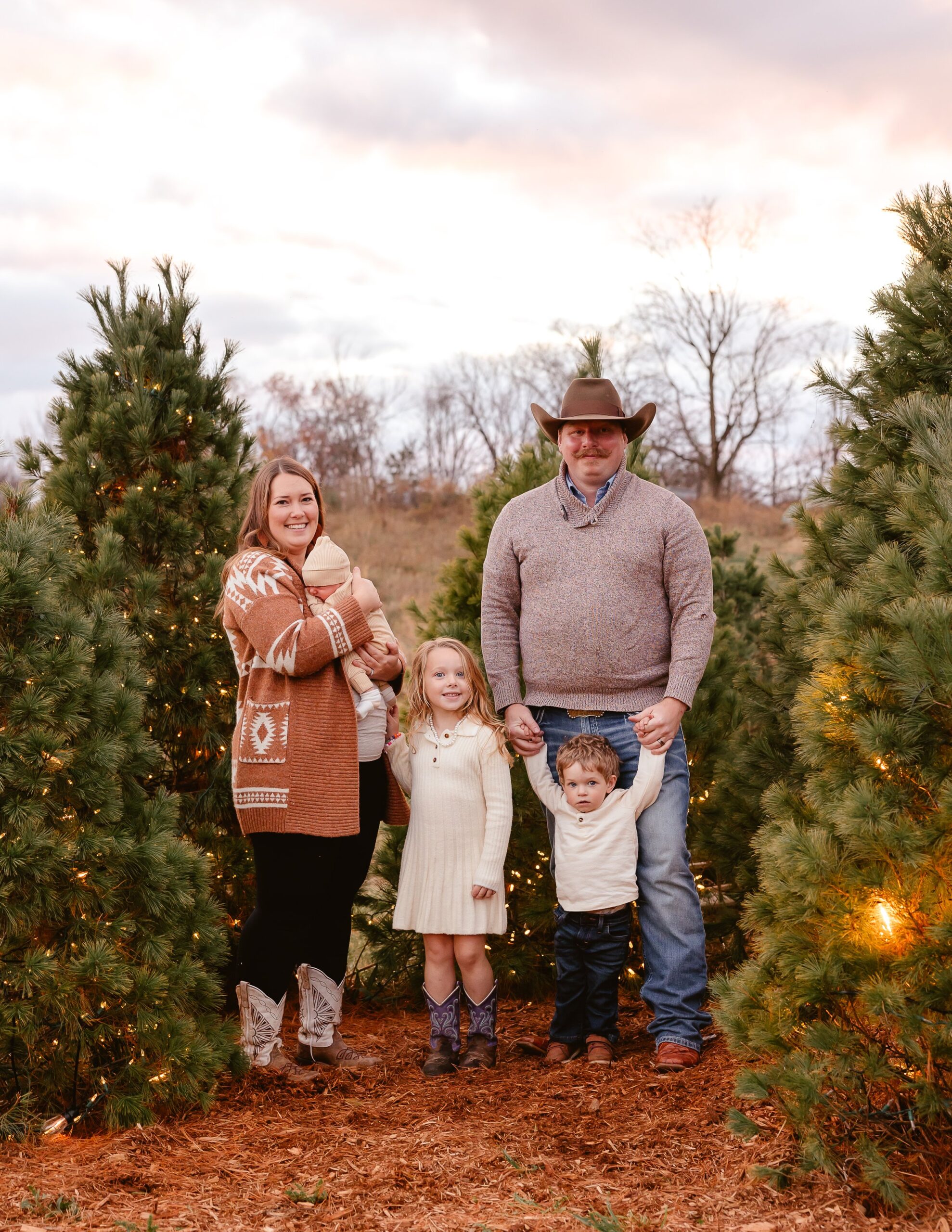 Family portrait in a Christmas tree farm, featuring a mother holding a baby, a father with a cowboy hat, a young girl in a white dress, and a little boy in a cream sweater. The scene is decorated with evergreen trees and warm lights, set against a cloudy sky.