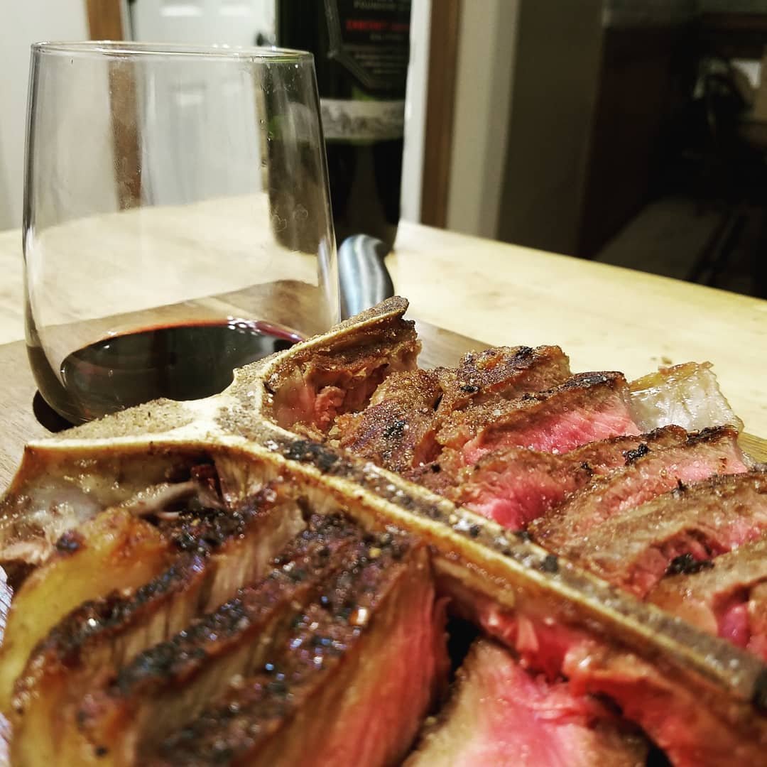 Close-up of a perfectly cooked T-bone steak, showcasing juicy pink meat with a charred exterior, served on a wooden board alongside a glass of red wine. A wine bottle is visible in the background, enhancing the gourmet dining experience.