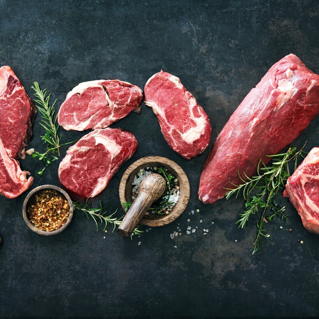 Assorted cuts of fresh beef, including ribeye and sirloin, arranged on a dark surface with sprigs of rosemary and bowls of spices, highlighting the quality and preparation for cooking.