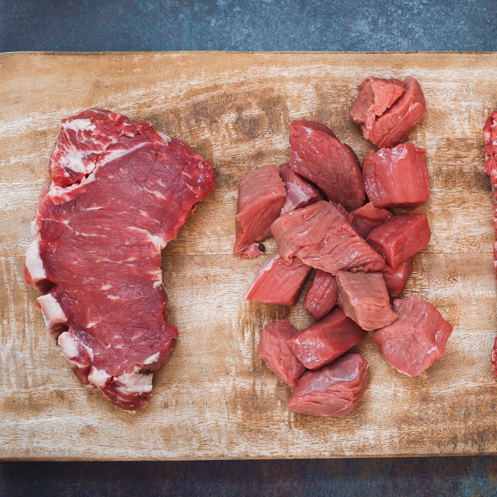 Fresh cuts of beef on a wooden cutting board, featuring a large ribeye steak on the left and neatly cubed pieces of beef in the center, ideal for cooking and meal preparation.