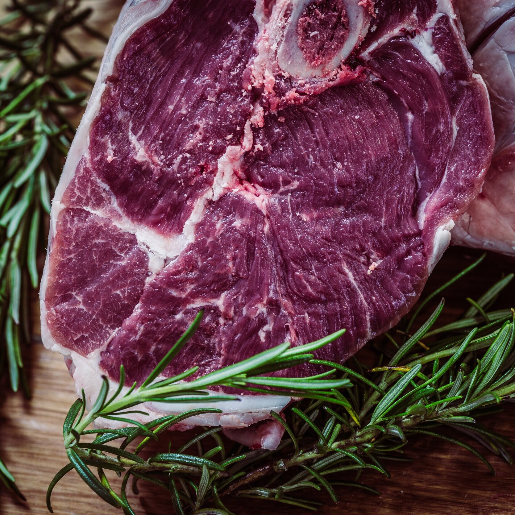 Close-up of a raw cut of meat, showcasing its marbling and texture, surrounded by fresh rosemary on a wooden surface, ideal for culinary uses and recipes.