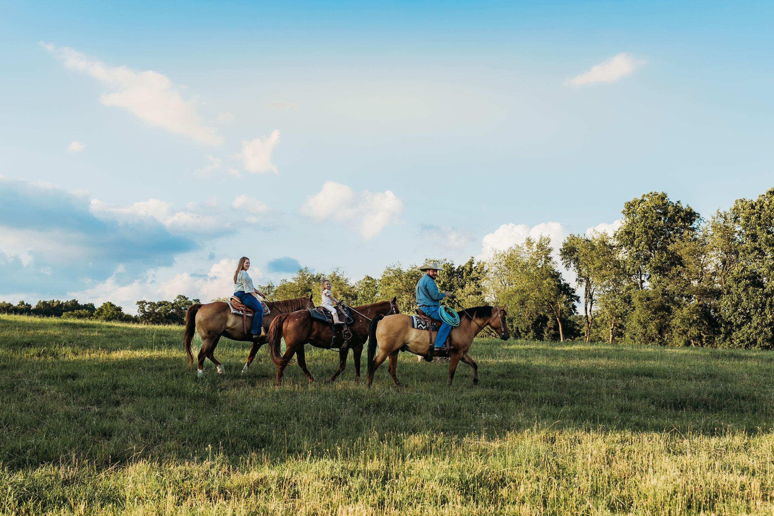 A group of people riding horses through a green field under a blue sky with clouds, showcasing a scenic outdoor equestrian experience.